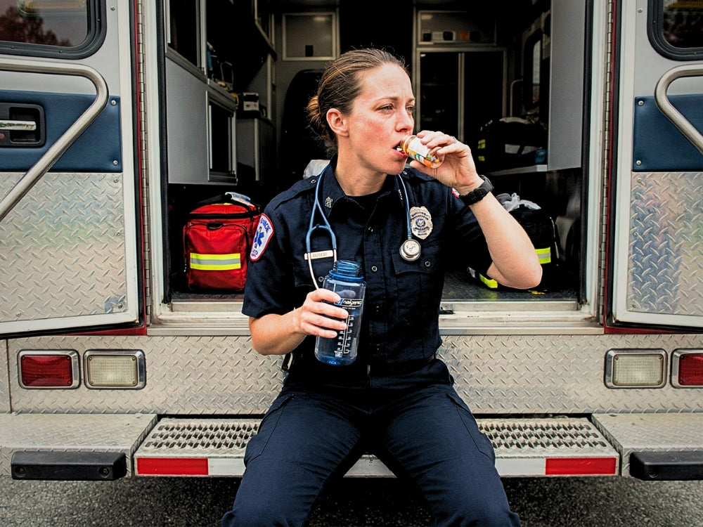 Person in an EMS uniform eating a sandwich in front of an ambulance.