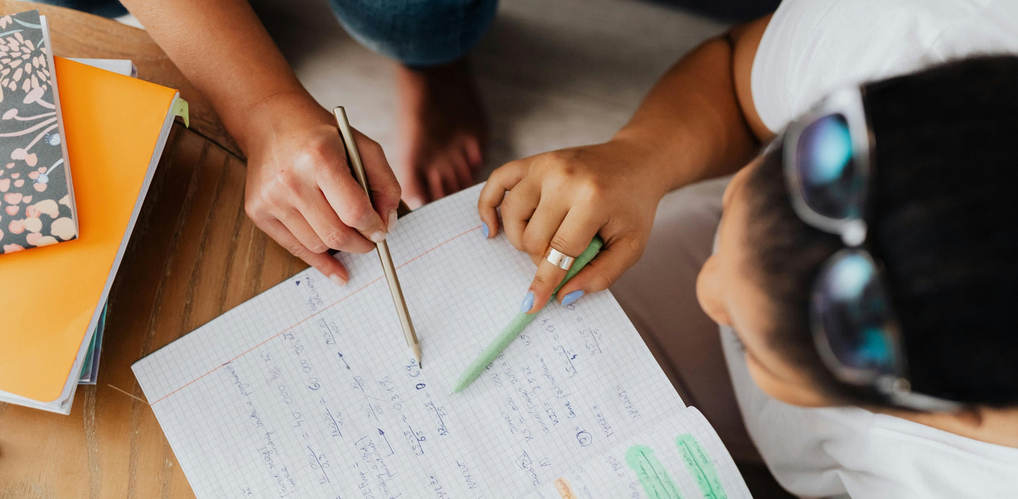 Two people working on a notebook with pens and notebooks on a table.